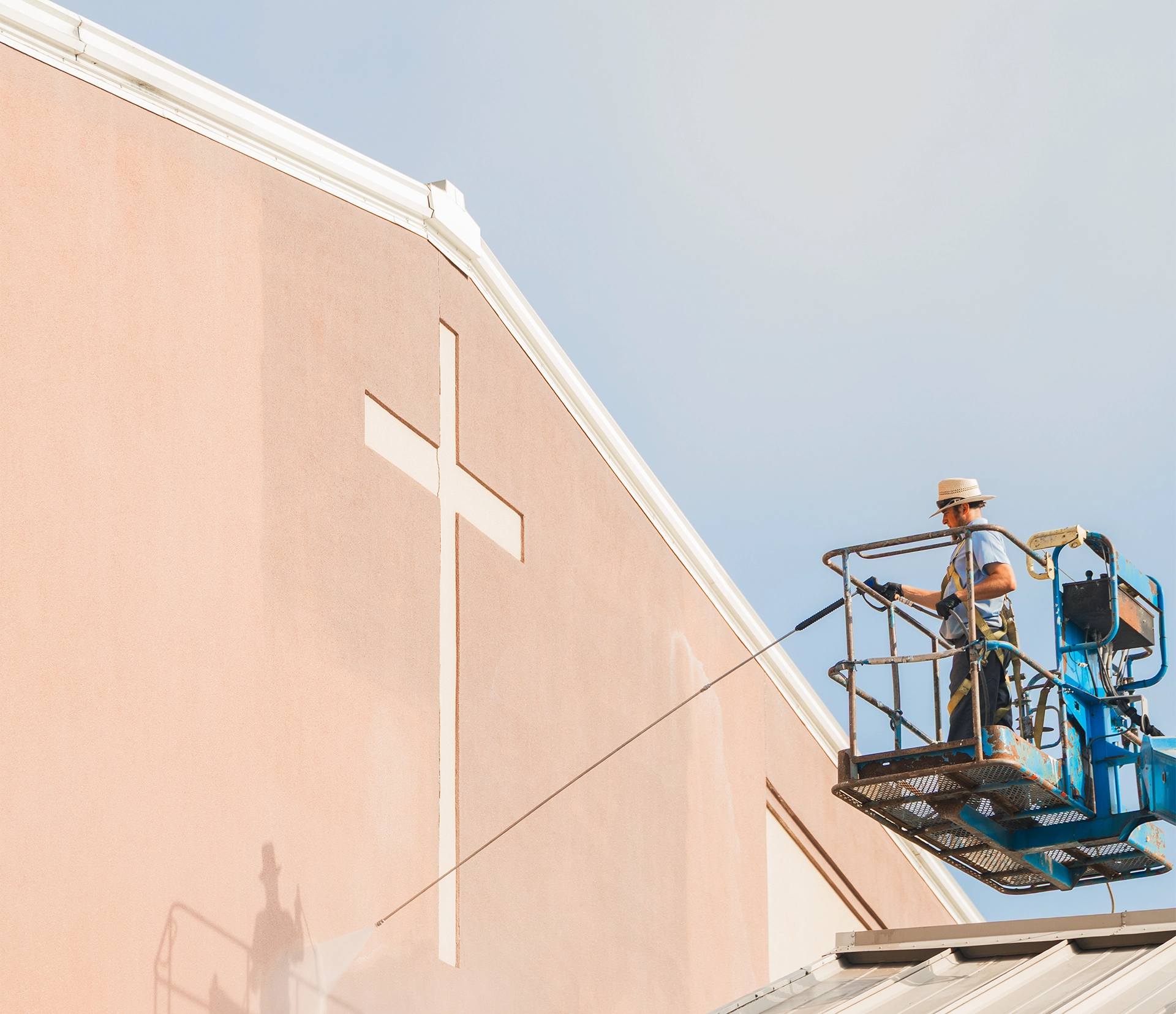 Man power washing a high church building wall with a cross cutout, elevated on a blue lift.