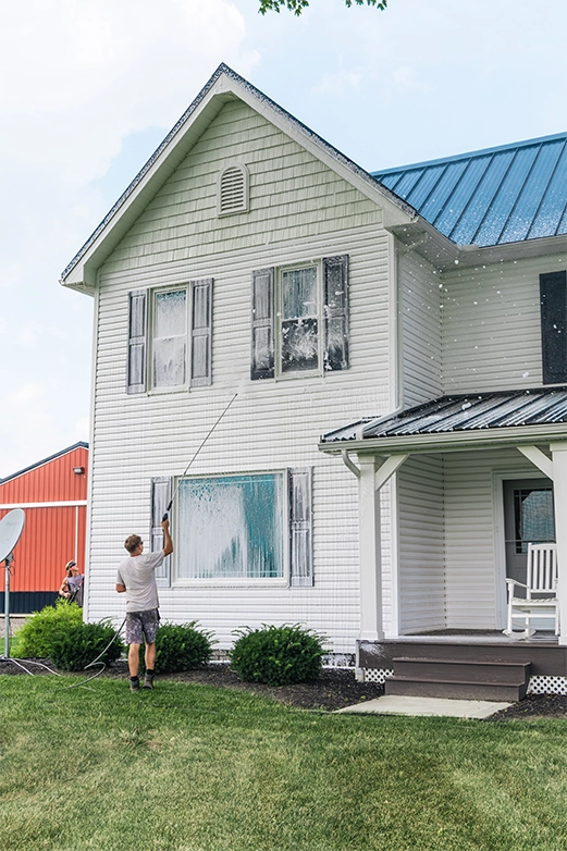 Man on ladder pressure washing a dark shingled roof with a spray nozzle.