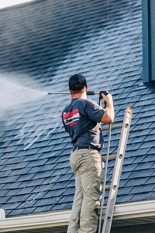 Man on ladder pressure washing a dark shingled roof with a spray nozzle.