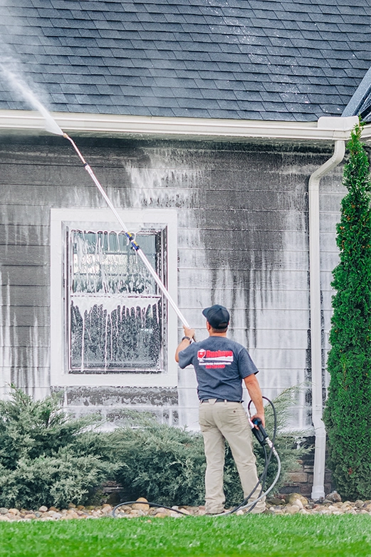Man on ladder pressure washing a dark shingled roof with a spray nozzle.