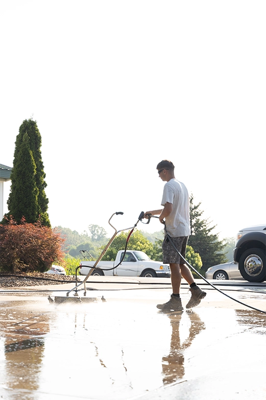 Man on ladder pressure washing a dark shingled roof with a spray nozzle.