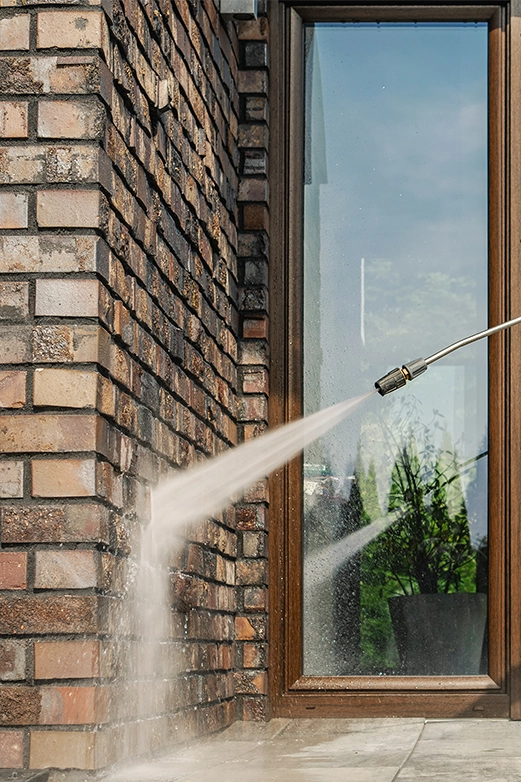 Man on ladder pressure washing a dark shingled roof with a spray nozzle.