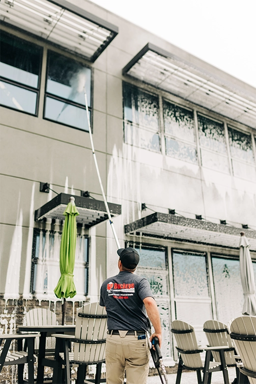 Man on ladder pressure washing a dark shingled roof with a spray nozzle.