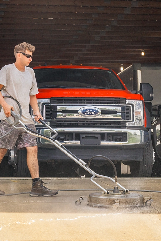 Man on ladder pressure washing a dark shingled roof with a spray nozzle.