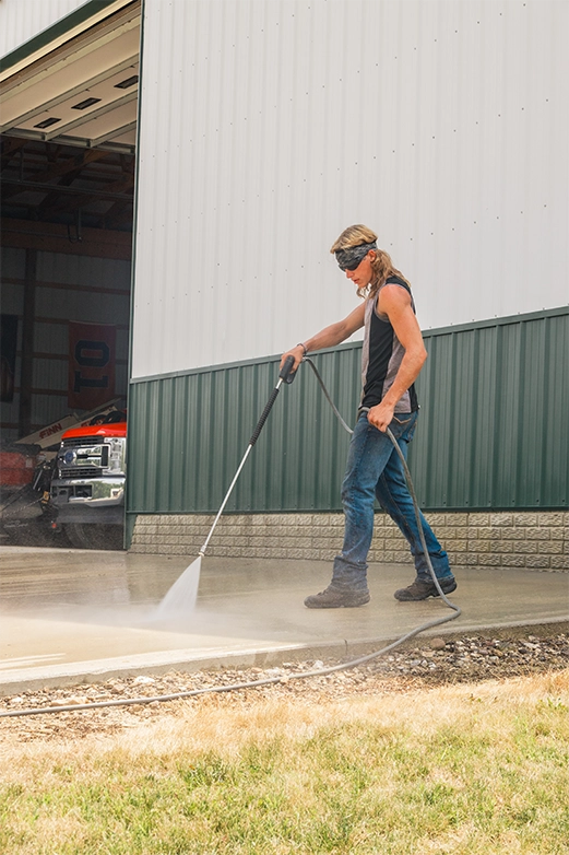 Man on ladder pressure washing a dark shingled roof with a spray nozzle.