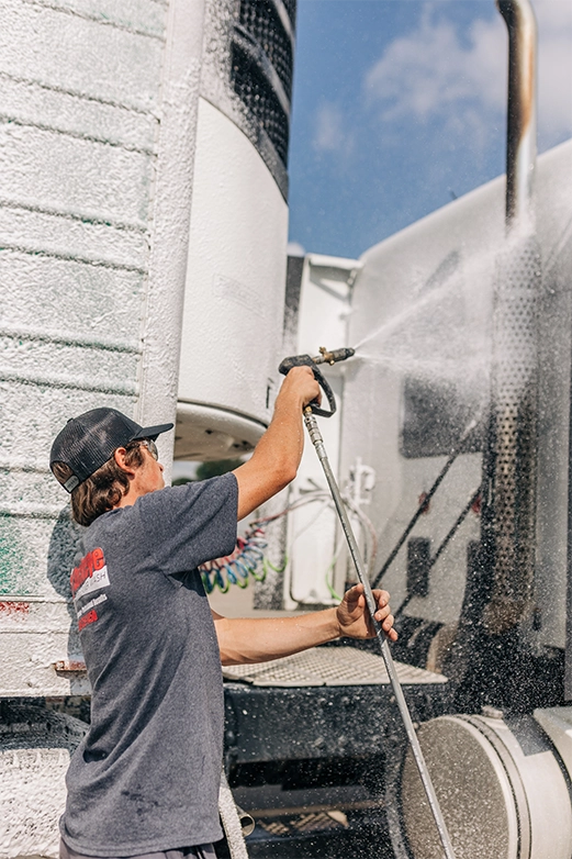 Man on ladder pressure washing a dark shingled roof with a spray nozzle.