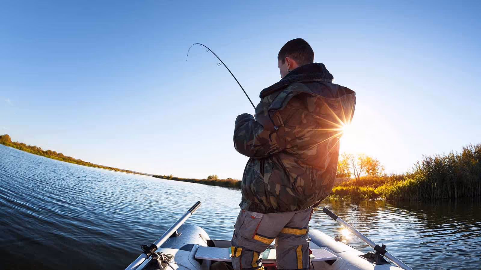 Man in jacket fishing in a calm lake with a sunset in front of him