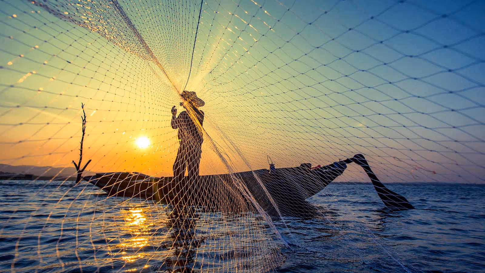 Man wearing a hat casting a fishing net into calm waters at sunset, capturing traditional fishing techniques and serene coastal scenery.