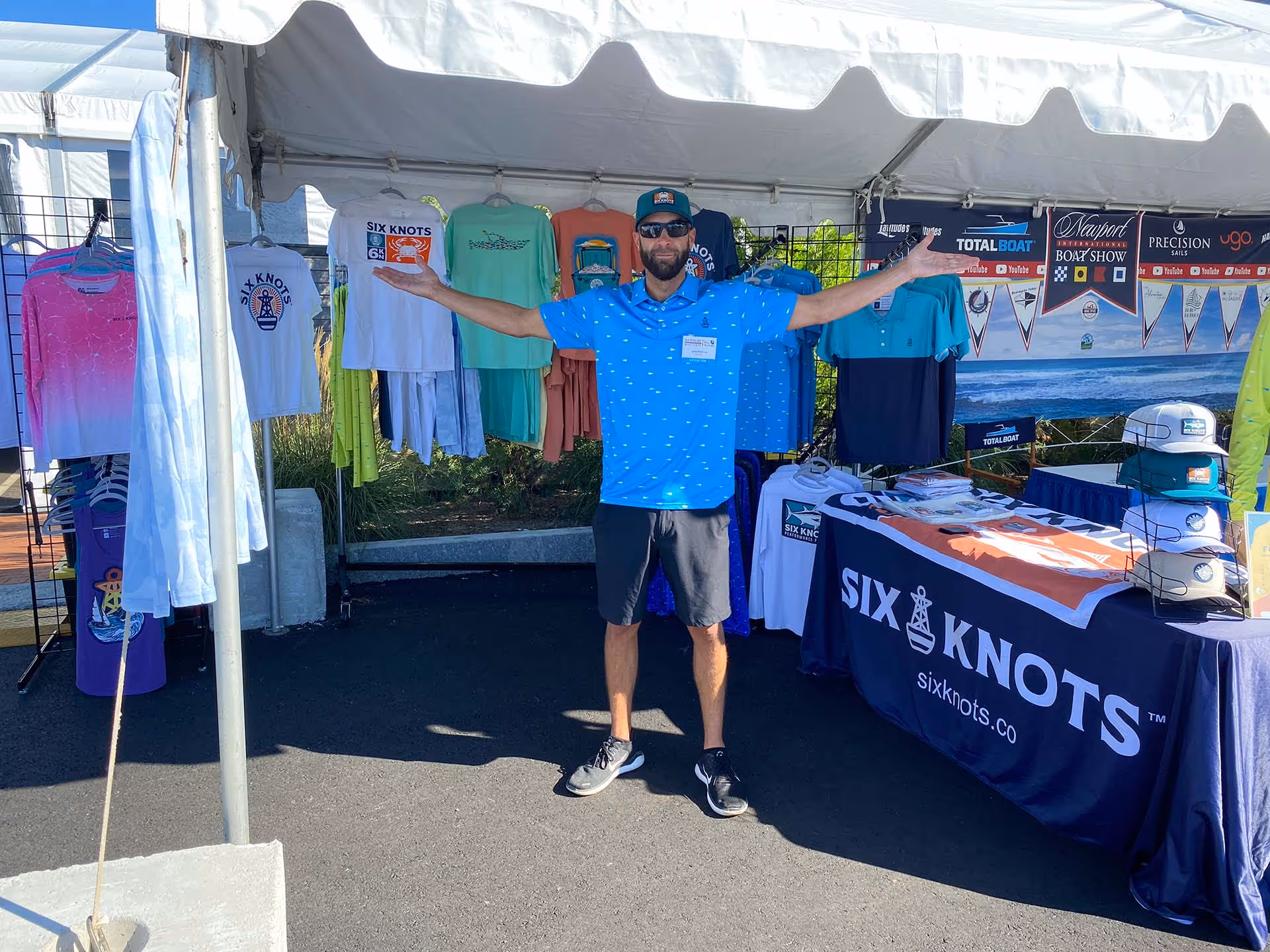 John from Six Knots proudly gestures with open arms in front of the Six Knots booth at the Newport International Boat Show