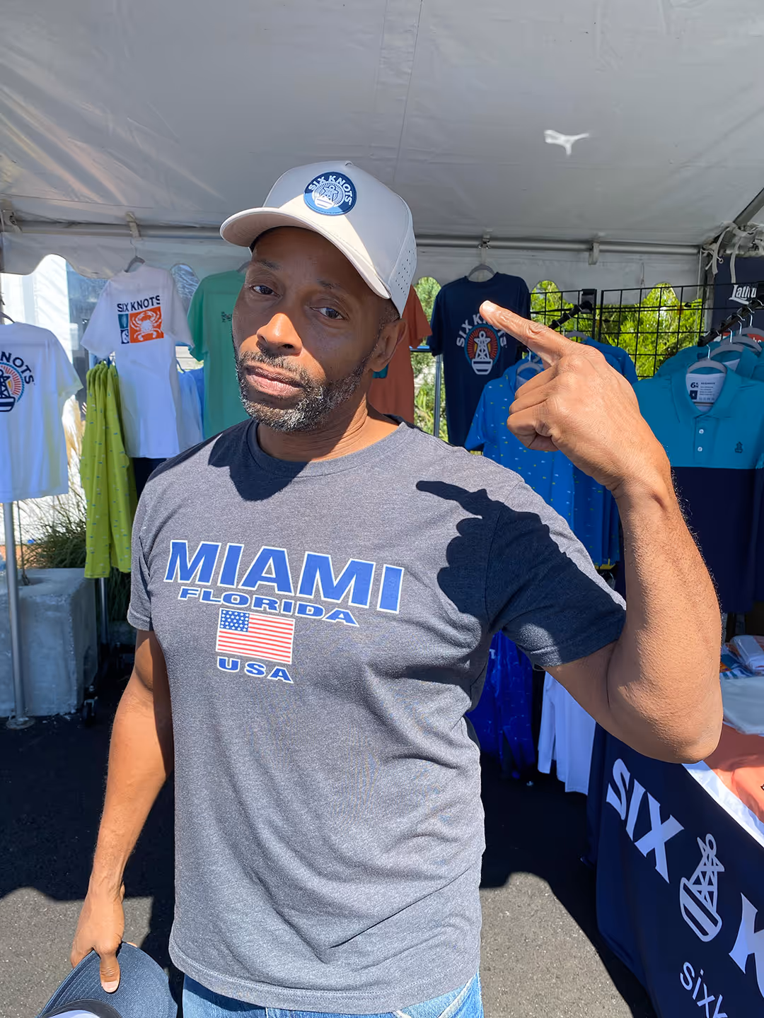 Man proudly points to his new Six Knots Captain hat in front of the booth at the Newport International Boat Show