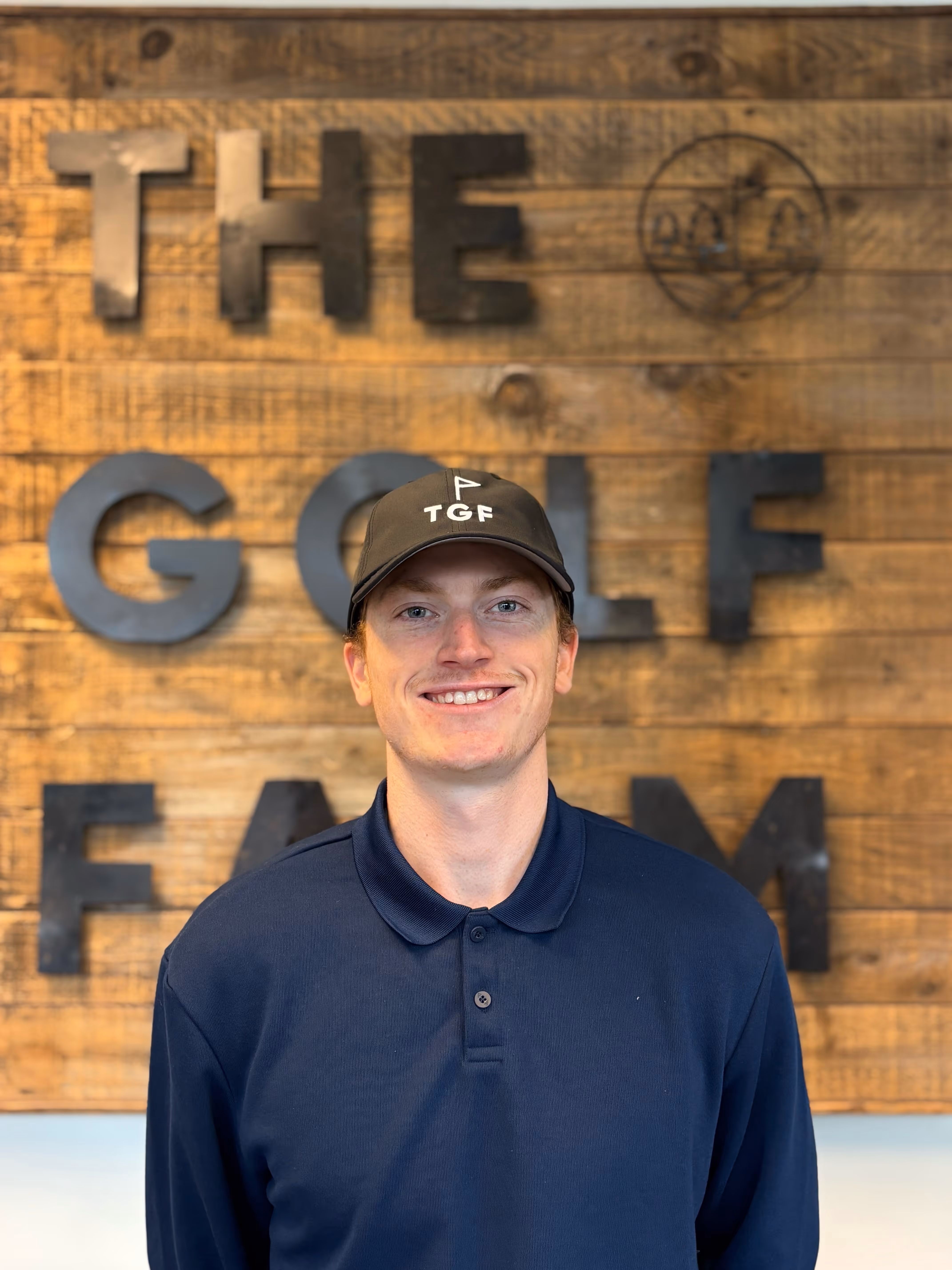 Smiling young man wearing a black TGF cap and navy blue polo shirt standing in front of a wooden wall with 'THE GOLF FARM' in black letters.