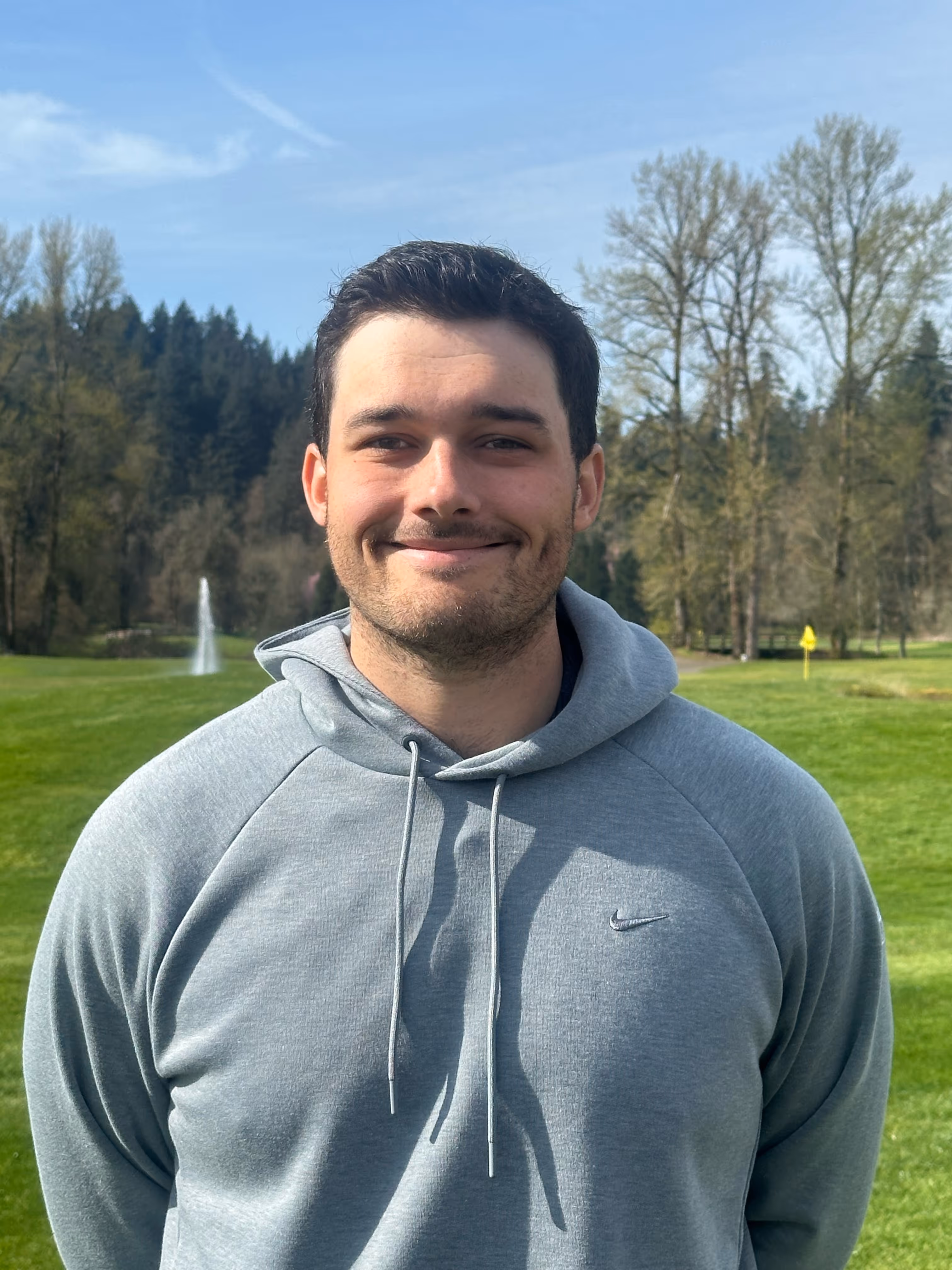 Smiling man in a grey Nike hoodie standing outdoors on a sunny day with green grass and trees in the background.