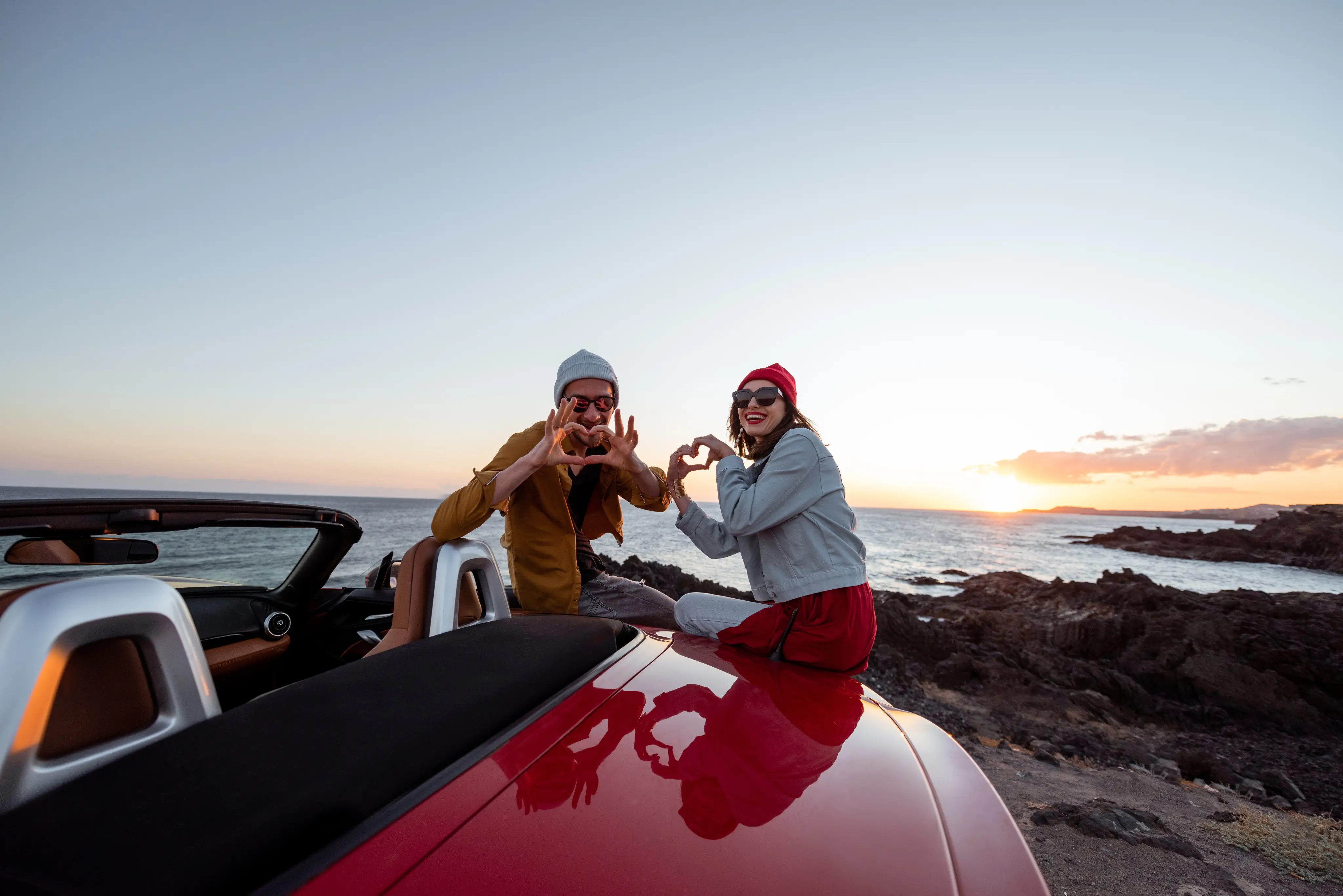 A couple sits on the edge of a parked car, making playful heart gestures with their hands.