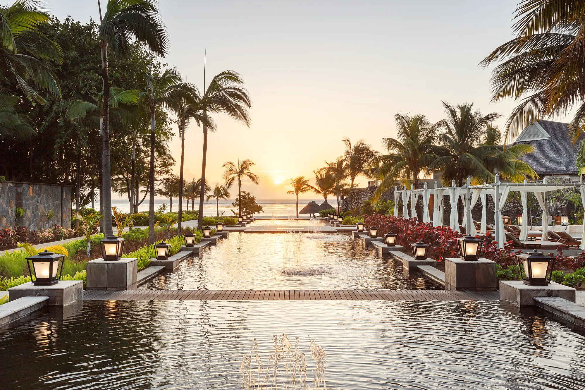 Lång reflekterande vattenspegel kantad av lyktor och palmer ner mot havet i solnedgången på The St. Regis Le Morne på Mauritius