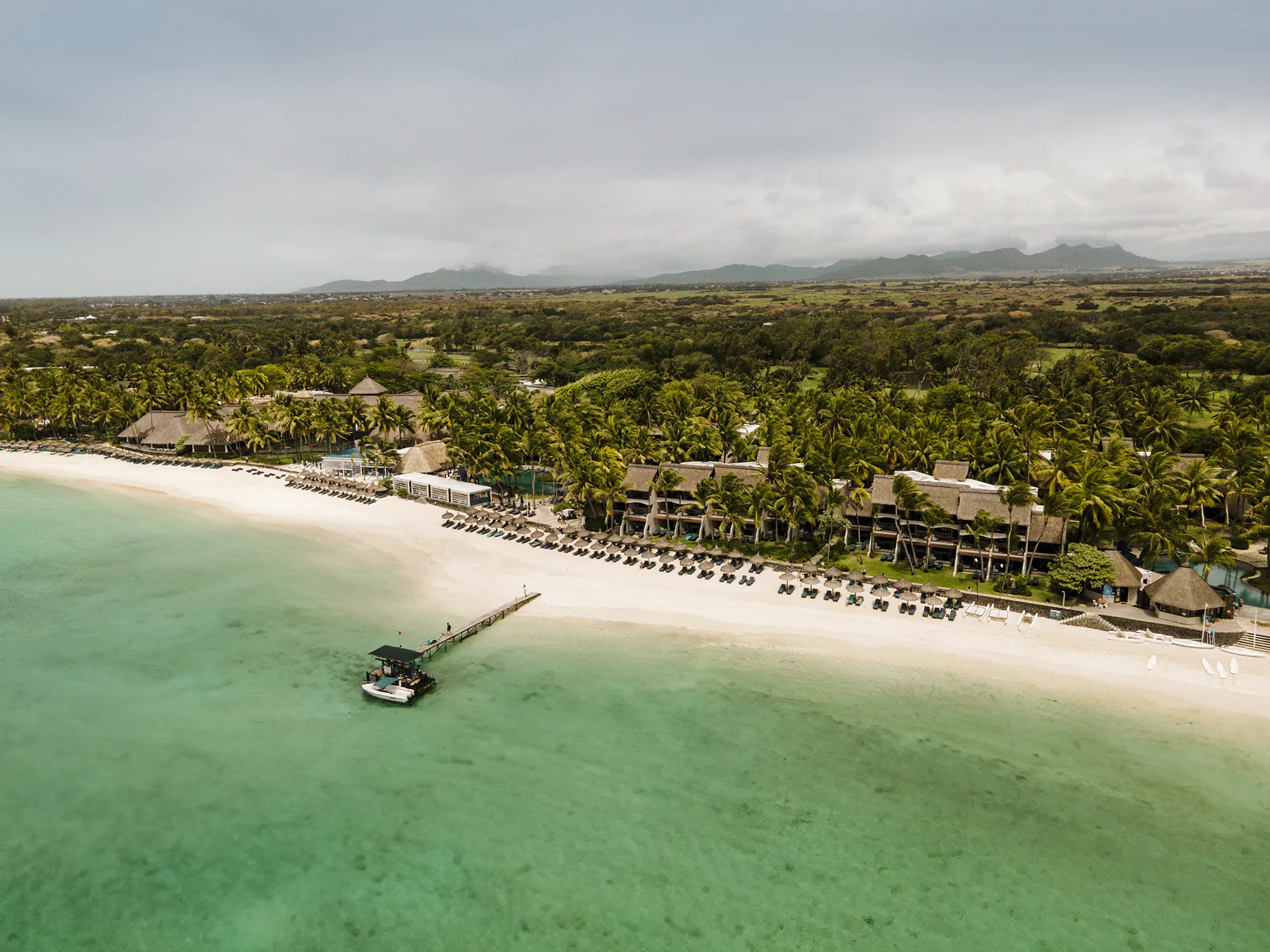 Flygbild över resortet längs en lång vit sandstrand med en brygga ut i lagunen och berg i bakgrunden