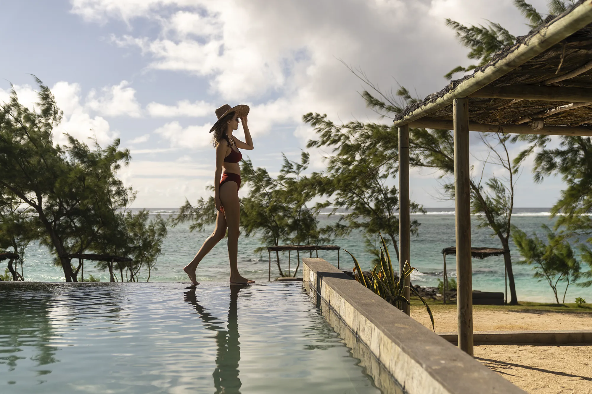 Kvinna i solhatt går längs kanten på en infinitypool med havet bakom