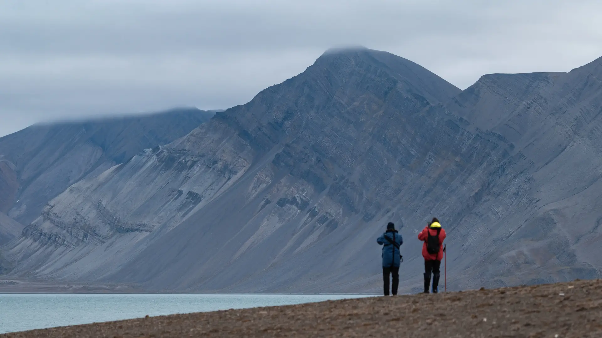 Miljöbild från Svalbard med natur och djurliv med två personer nära hav och framför berg är i fokus.