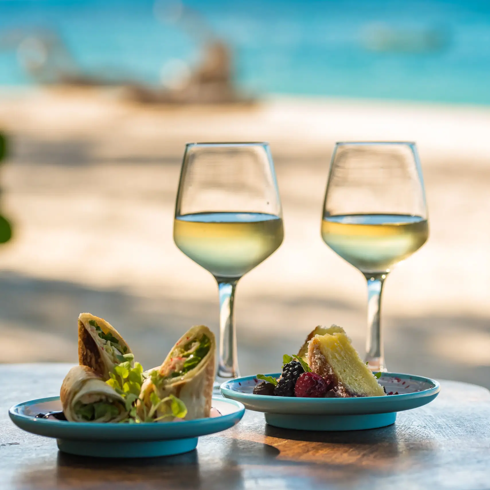 Två vinglas och mat i förgrunden, och en strand på Seychellerna i bakgrunden.