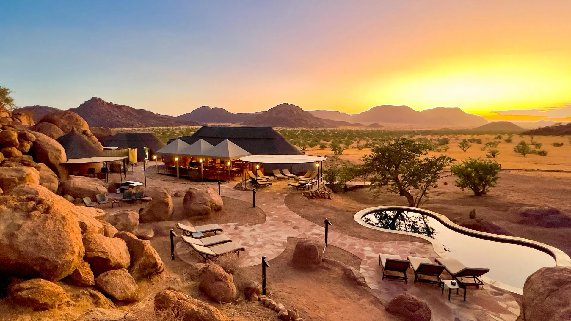     Lyxigt ökenresort i Namibia med pool, stenformationer och vidsträckt utsikt över landskapet i solnedgång.