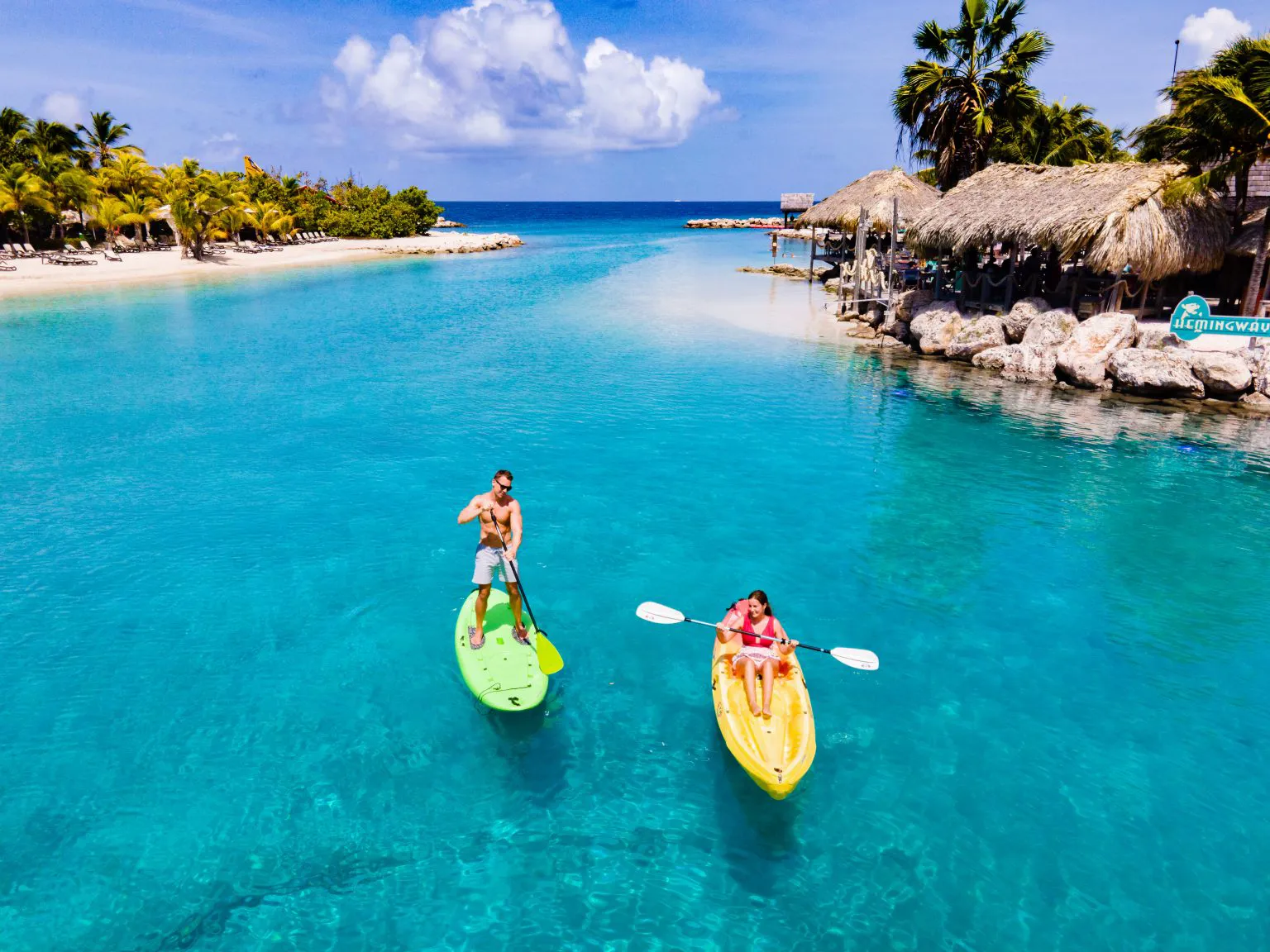 Personer paddlar SUP och kajak i turkost vatten vid tropisk strand och resort på Curaçao.