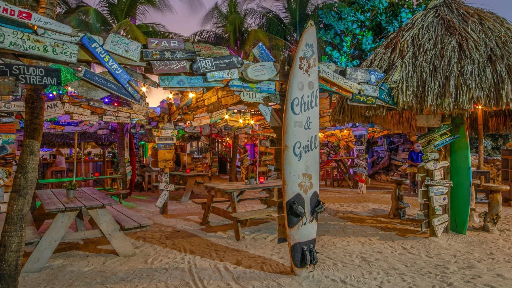 Färgstark strandbar på Curaçao med surfbrädor, palmtak och skyltar i tropisk kvällsmiljö vid stranden.