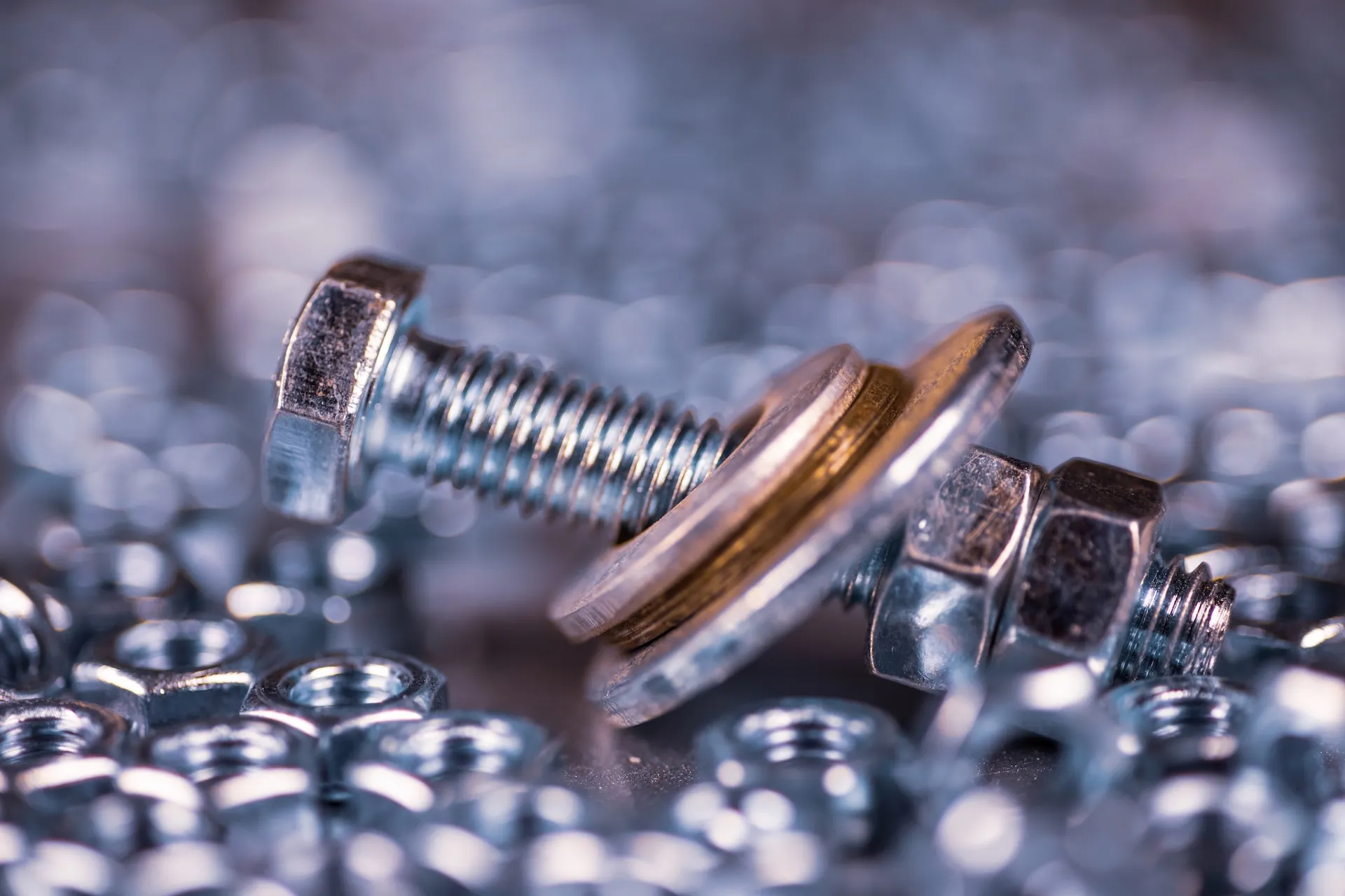 Close-up of metal bolt with two washers and a nut surrounded by scattered nuts on a reflective surface.
