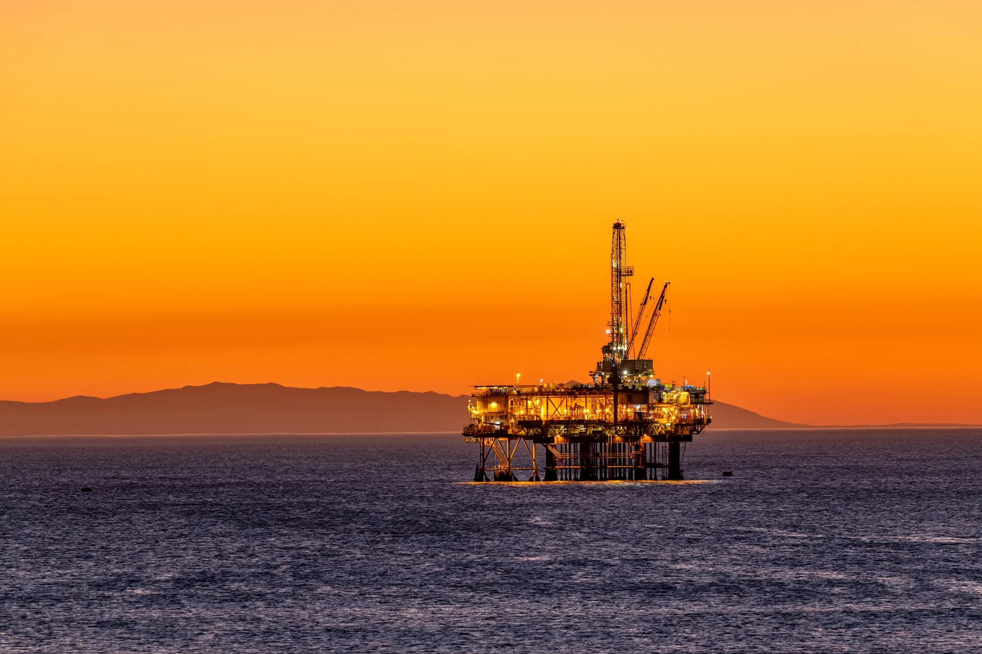 Illuminated offshore oil platform in the ocean at sunset with mountains in the background.