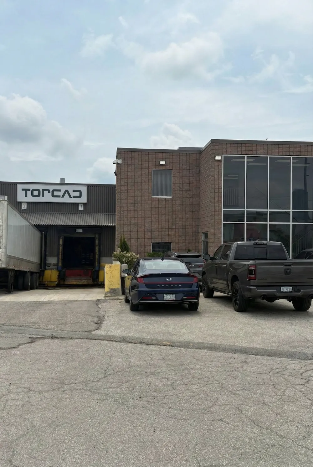 Front view of Torcad building with a large glass window, delivery dock with a trailer, and three parked vehicles on an overcast day.