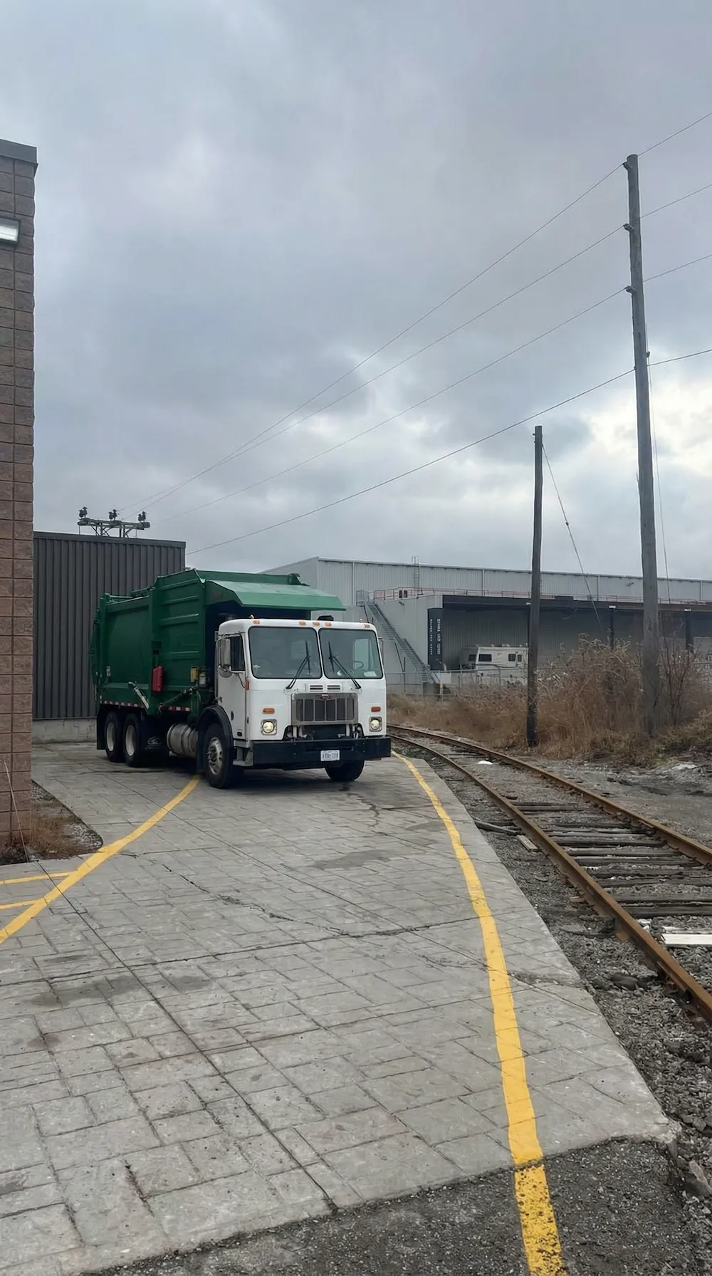 Blue and white garbage truck with a green leaf logo parked near an industrial building at sunset.