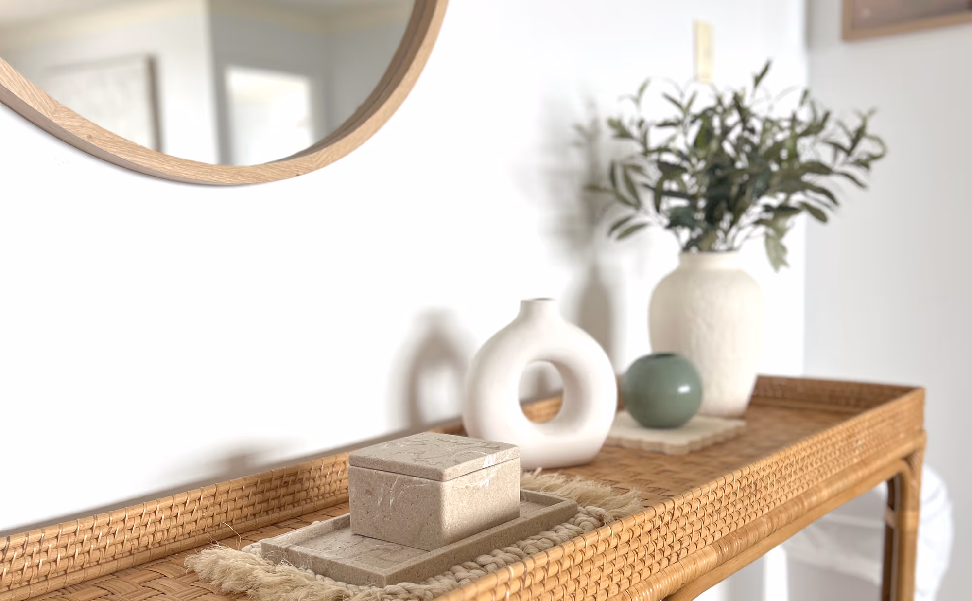 Wicker console table with decorative beige stone box, white circular vase, small green spherical vase, and a white vase with green foliage.