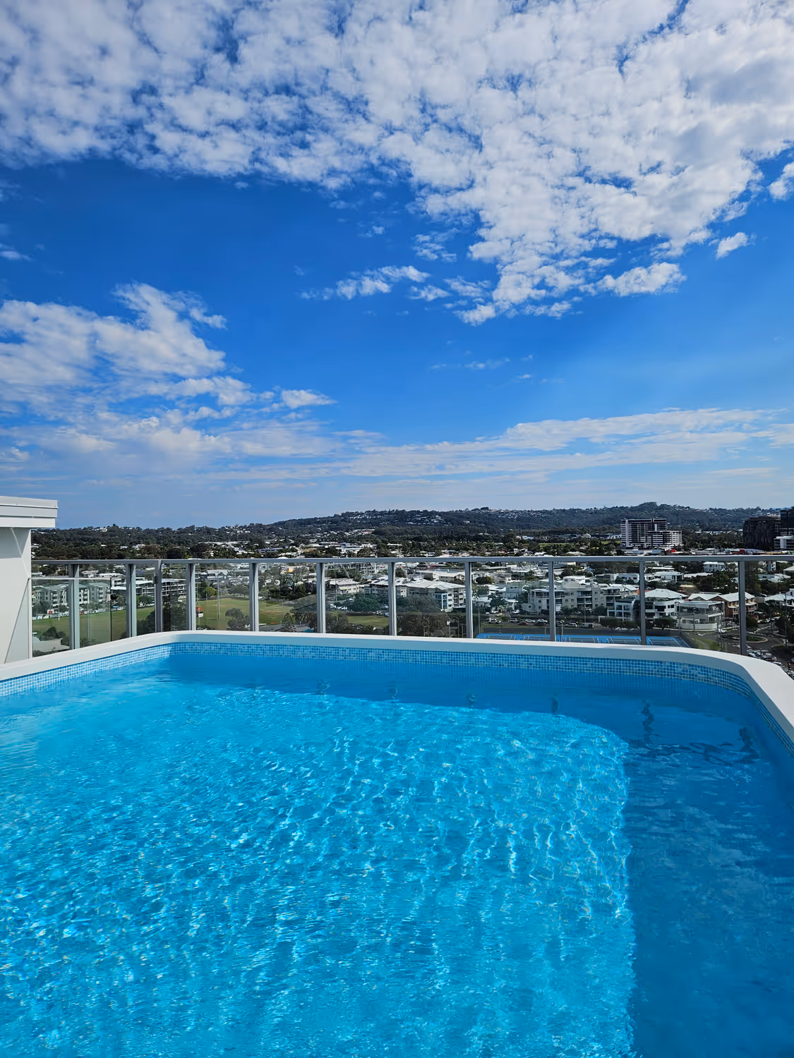 Clear blue rooftop pool overlooking a cityscape with hills under a partly cloudy sky.