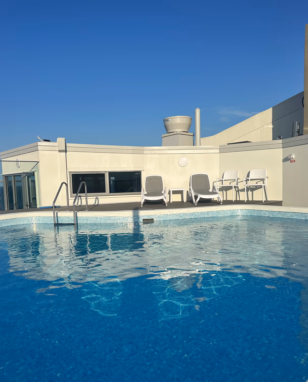 Clear rooftop swimming pool with two white lounge chairs, two white chairs, and a small table under a blue sky.