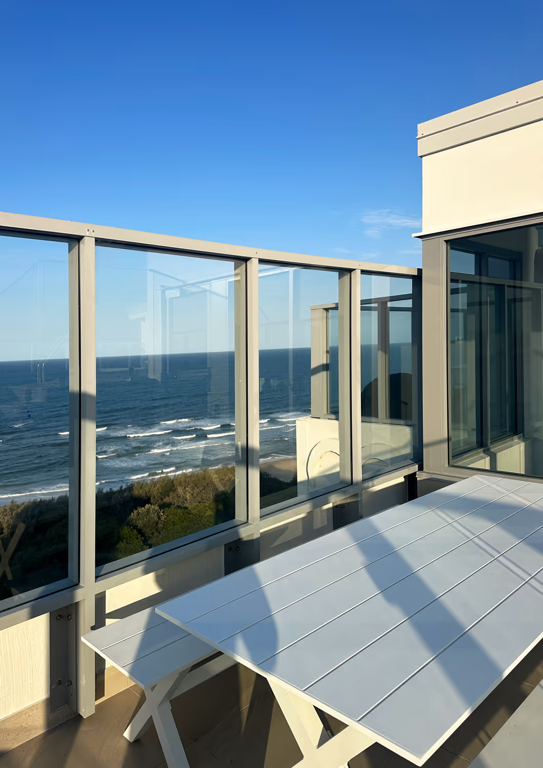 Balcony with white table and bench overlooking ocean waves under clear blue sky.
