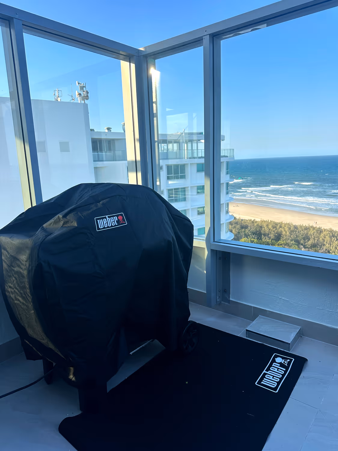 Covered Weber grill on a balcony with glass railing overlooking a beach and ocean under a clear blue sky.