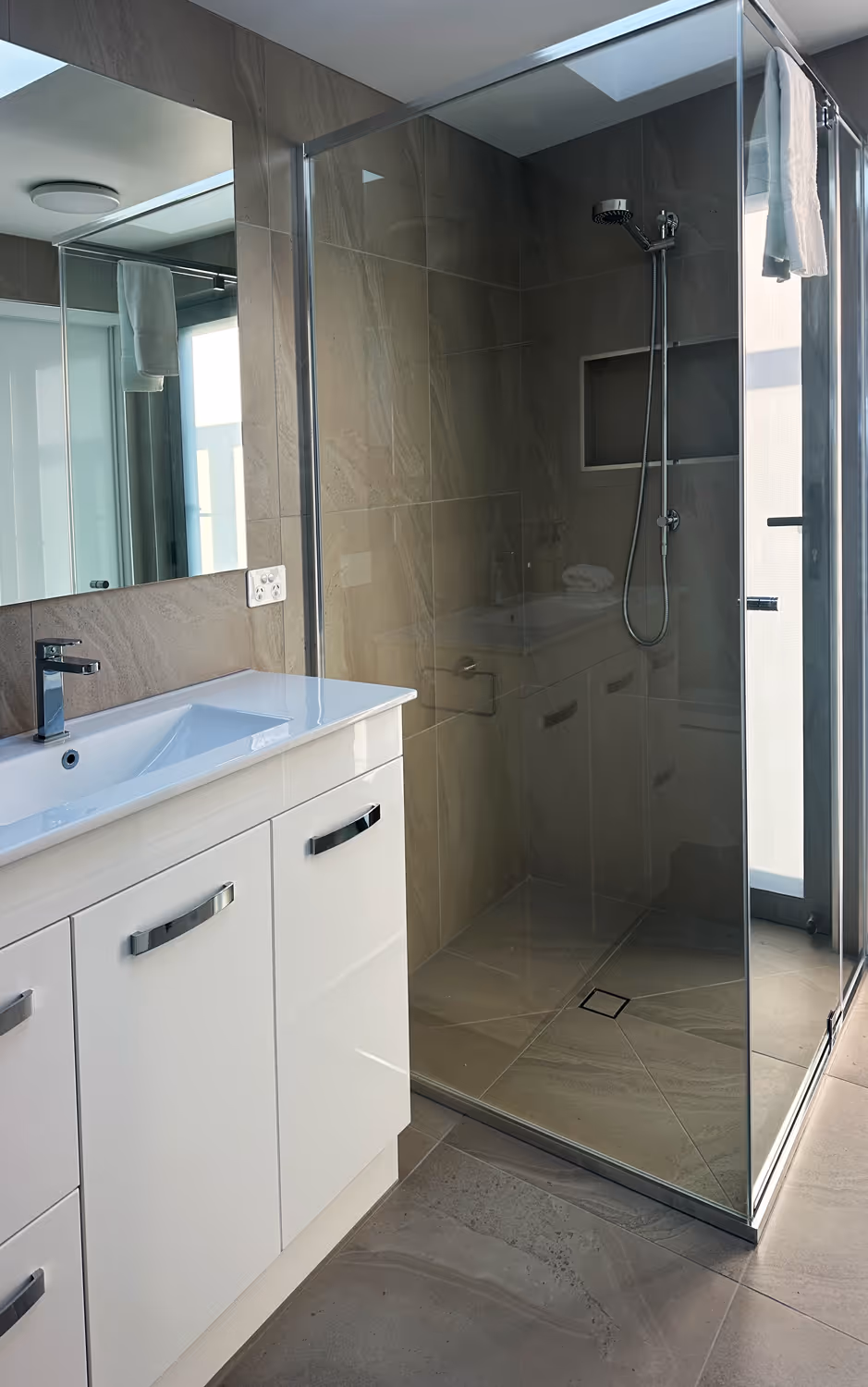 Modern bathroom with glass-enclosed shower, beige tile walls, and white sink vanity with chrome faucet.