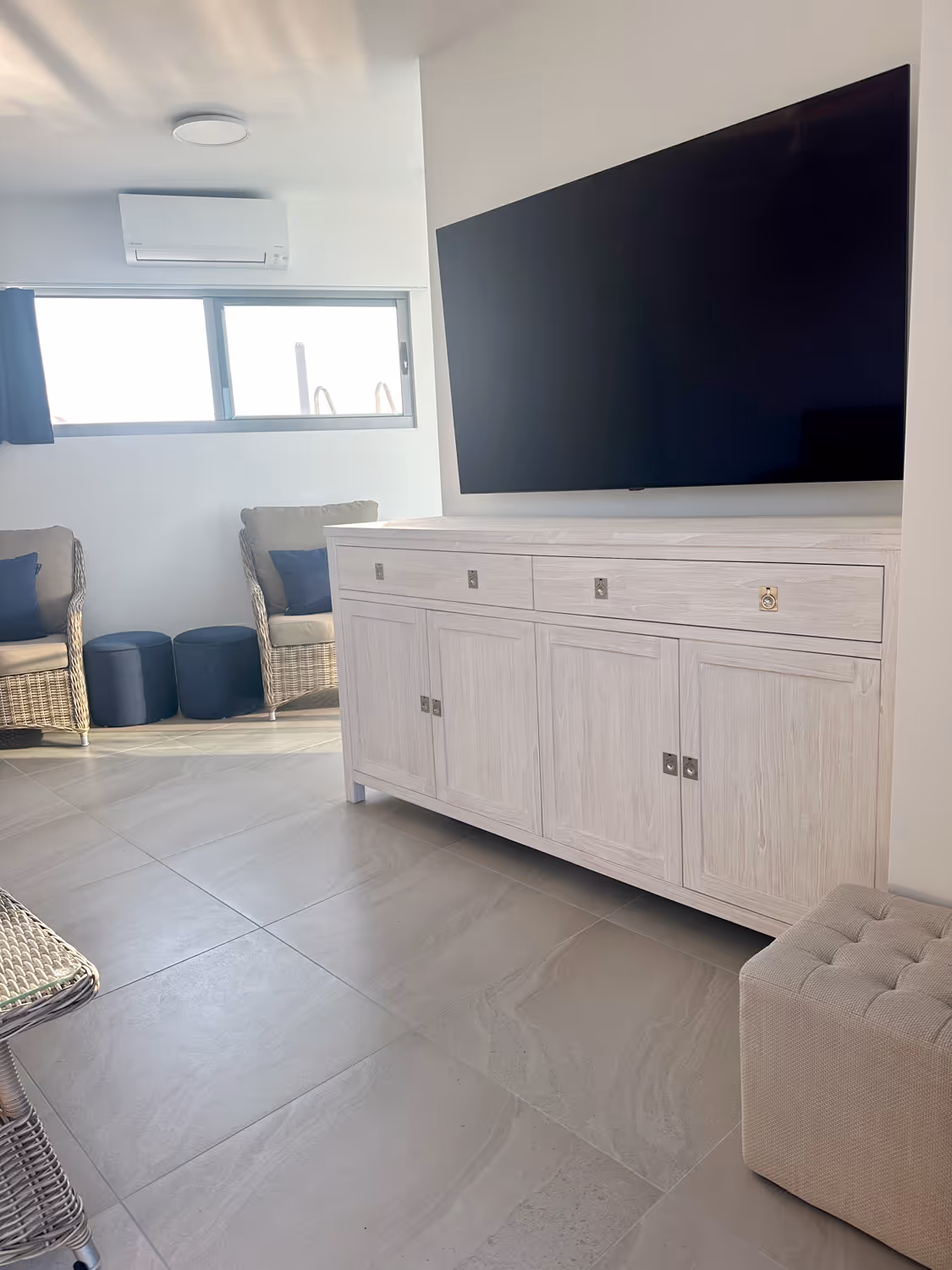 Modern living room corner with a large flat-screen TV mounted above a light wood cabinet, rattan chairs with navy cushions, two dark blue ottomans, and a beige tufted ottoman on tiled floor.