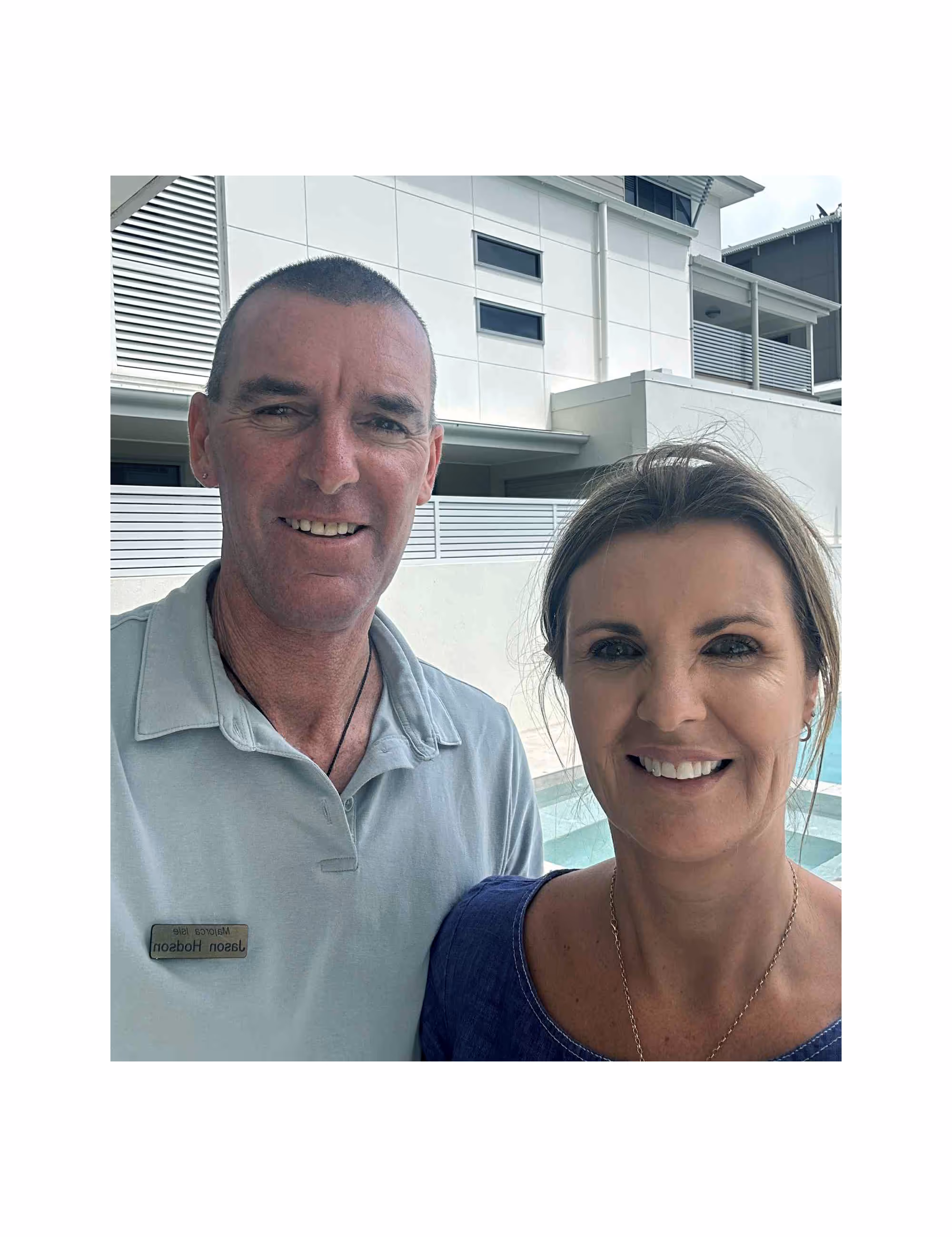 Smiling man and woman standing together outside near a building and pool, man wearing a name tag.