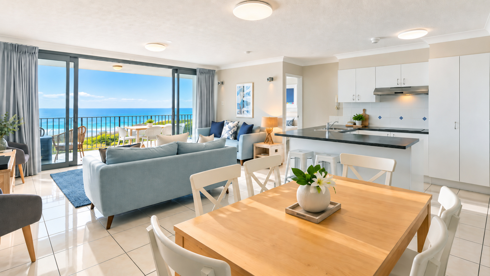 Bright open-plan living space with light blue couches, wooden dining table with white chairs, modern white kitchen with black countertops, and large sliding glass doors opening to a balcony overlooking a blue ocean and clear sky.