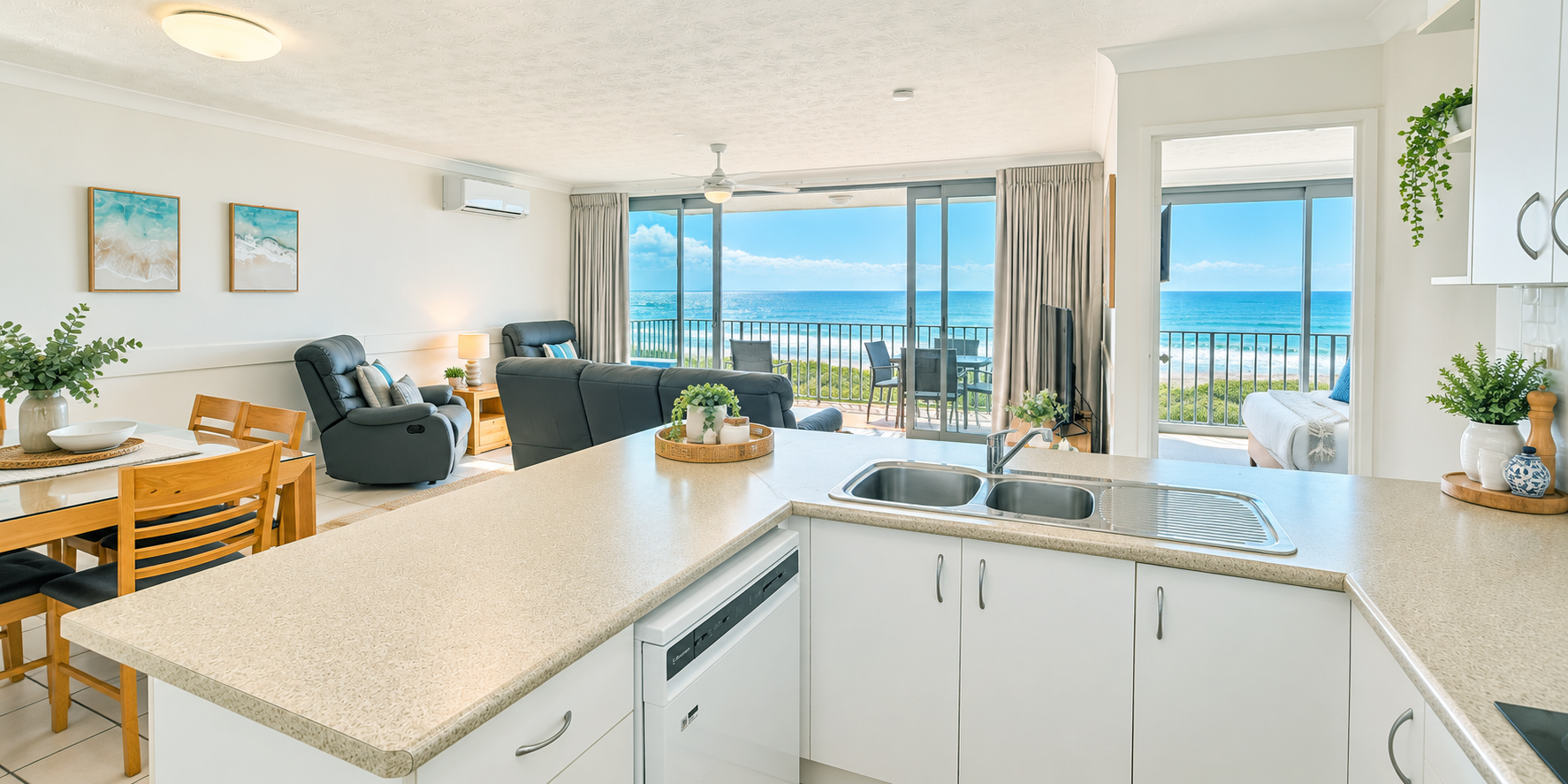 Bright open-plan living area with a kitchen counter in the foreground featuring a double sink and dishwasher, a dining table with wooden chairs, black leather armchairs and sofa, and sliding glass doors opening to a balcony with patio furniture and an ocean view under a partly cloudy sky.