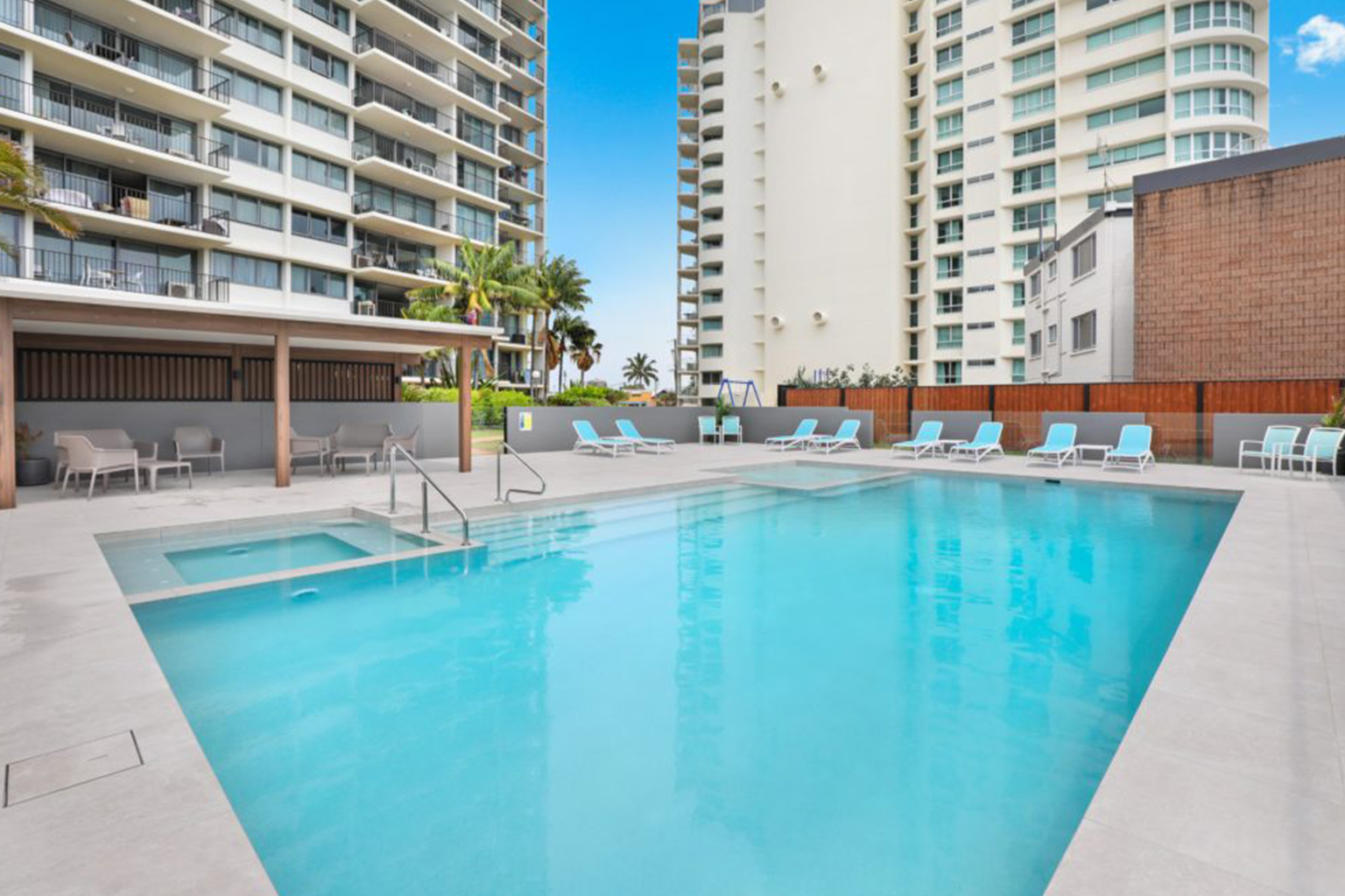 Clear blue swimming pool with a connected smaller hot tub, surrounded by light-colored tiles and lounge chairs with blue cushions, set between modern apartment buildings under a clear sky.
