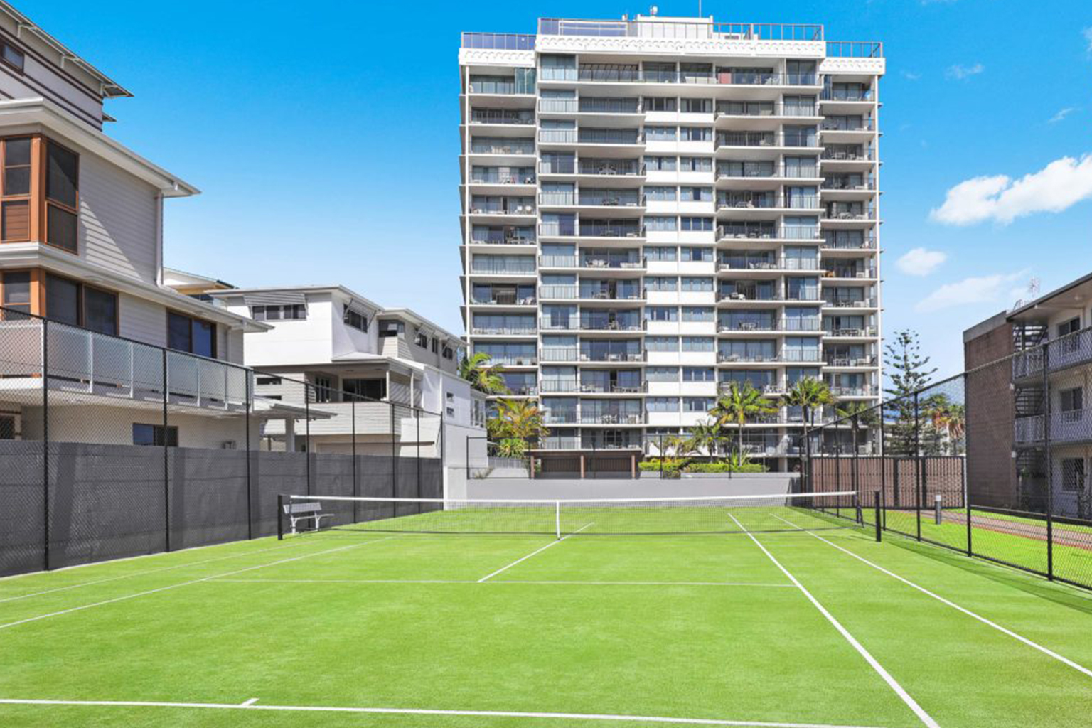 Outdoor grass tennis court with white lines and net, enclosed by black chain-link fence, surrounded by multi-story residential buildings under a clear blue sky.