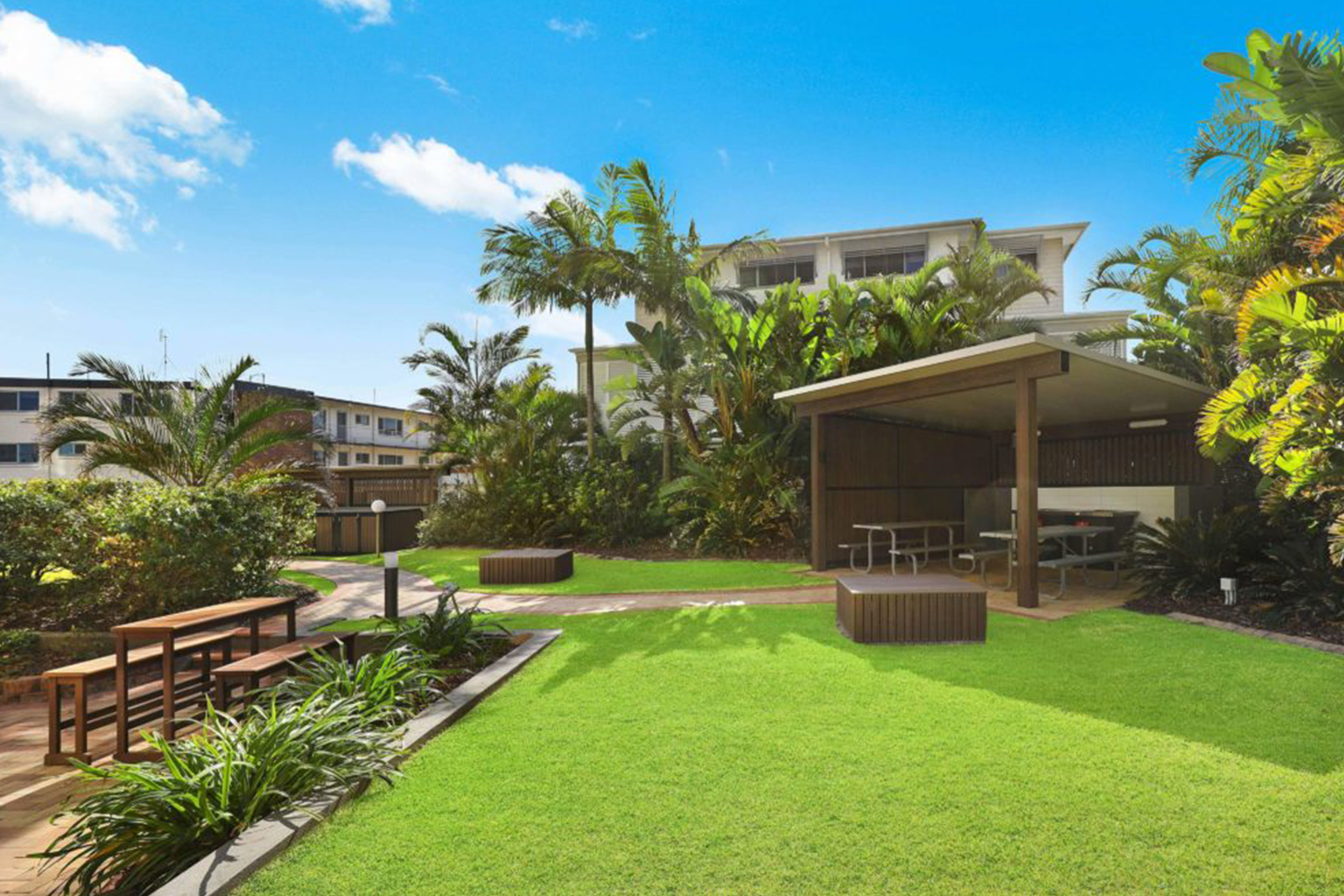 Outdoor area with well-maintained green lawn, wooden benches and picnic tables under a covered pavilion, surrounded by tropical plants and palm trees, with a clear blue sky overhead and residential buildings in the background.