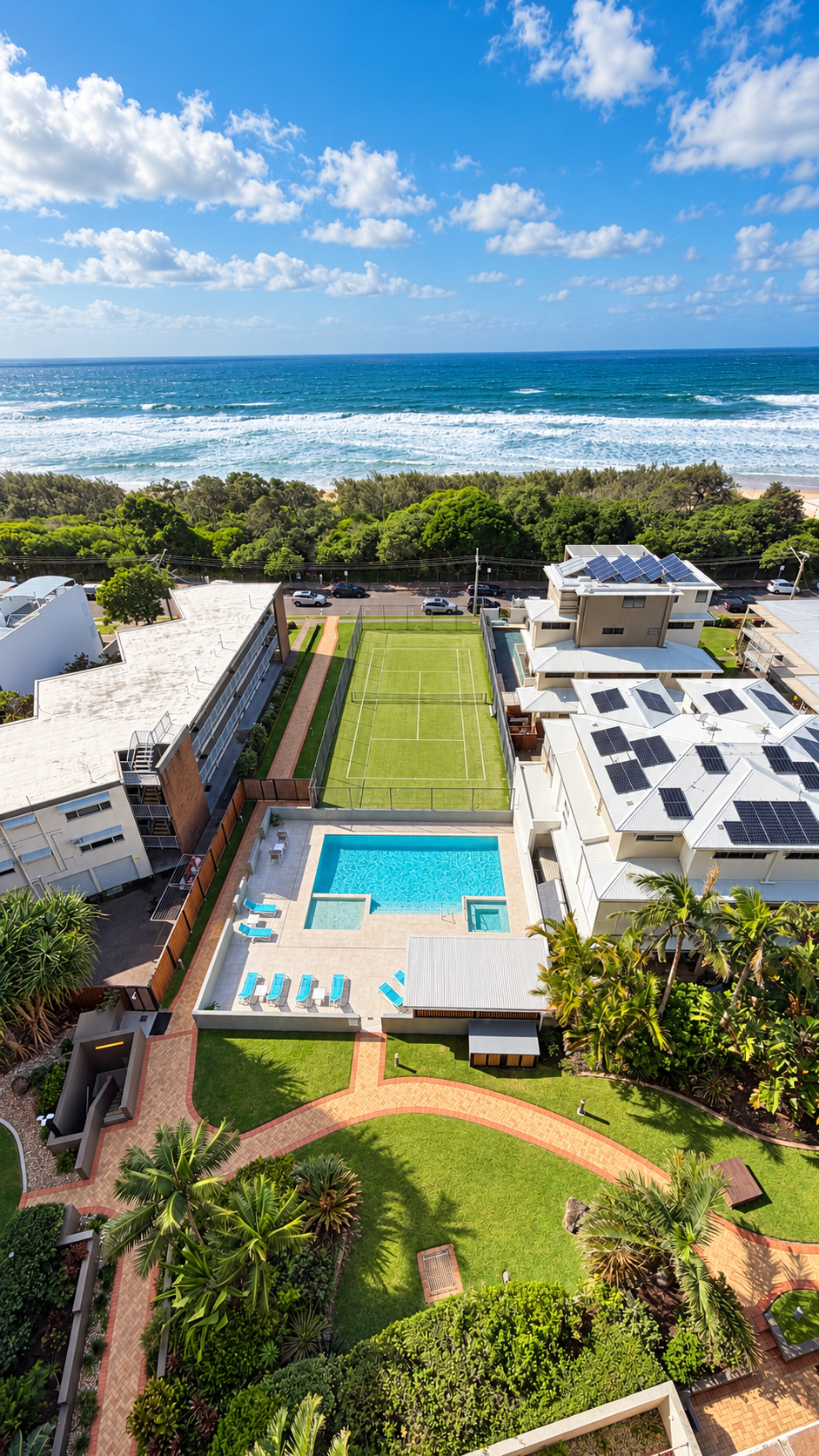 Aerial view of a resort area featuring a rectangular swimming pool with lounge chairs, a small tennis or paddle court with marked lines, lush green lawns with walking paths, and residential buildings with solar panels on the roofs, all near a road lined with trees and the ocean with waves in the background under a partly cloudy blue sky.