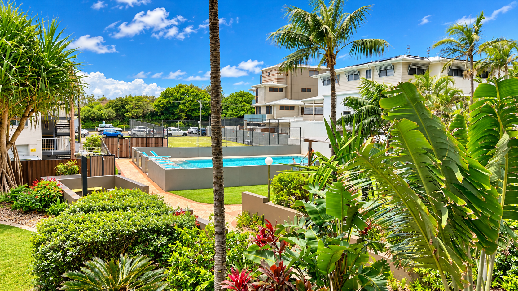 Outdoor view of a residential pool surrounded by a low gray wall and lounge chairs, with lush tropical plants and palm trees in the foreground and multi-story buildings behind, under a blue sky with scattered clouds.
