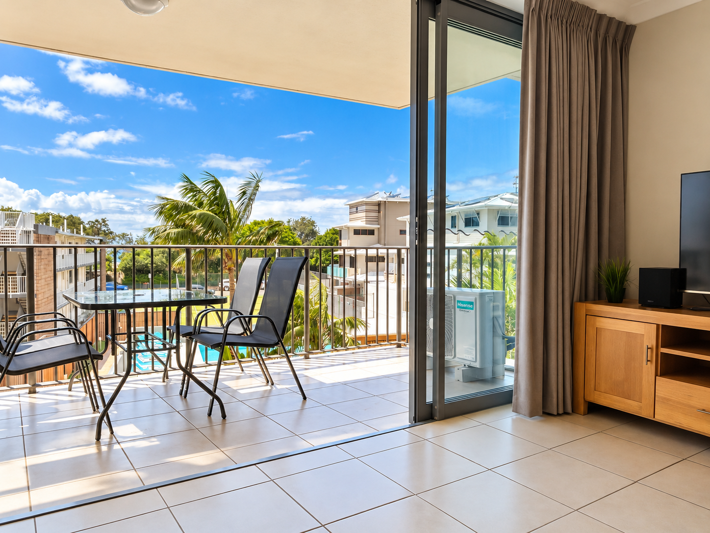 Balcony with glass-top table and black mesh chairs overlooking palm trees, buildings, and a clear blue sky, seen from a tiled interior living space with brown curtains and a wooden TV cabinet.