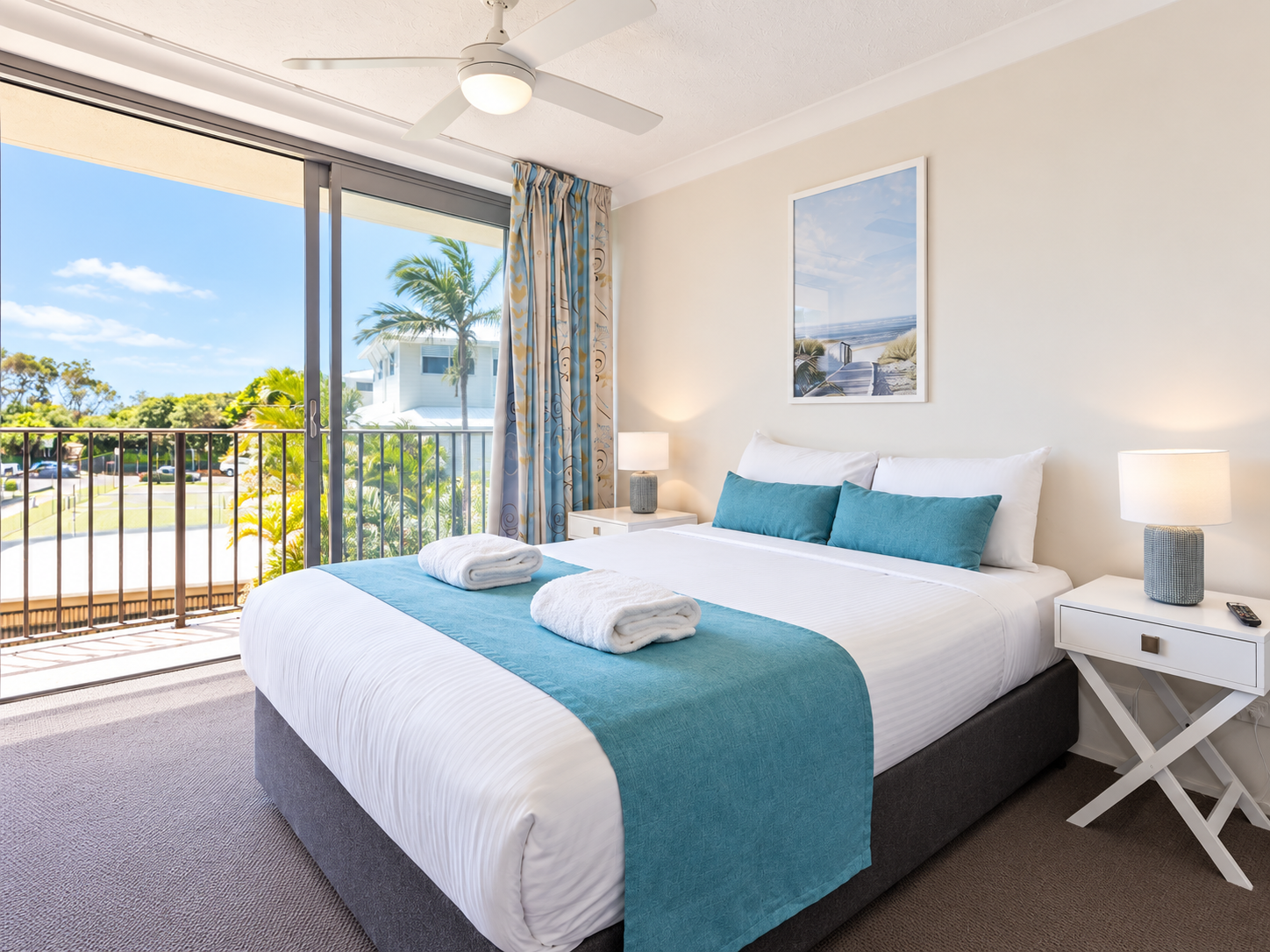 Bright master bedroom with a large bed dressed in white linens and teal accents, two white bedside tables with lamps, and a framed beach scene above the bed with a sliding glass door leading to a balcony showing palm trees and a clear blue sky.