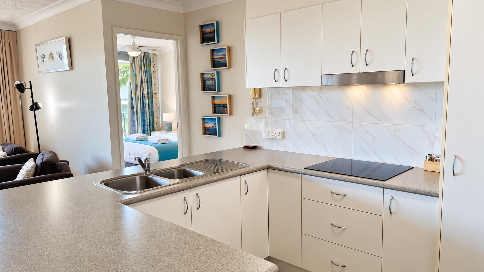 Modern kitchen with light beige countertops, double stainless steel sink, electric cooktop, and white cabinetry with silver handles; in the background, a bedroom with a bed, blue accents, and rolled towels is visible through an open doorway.