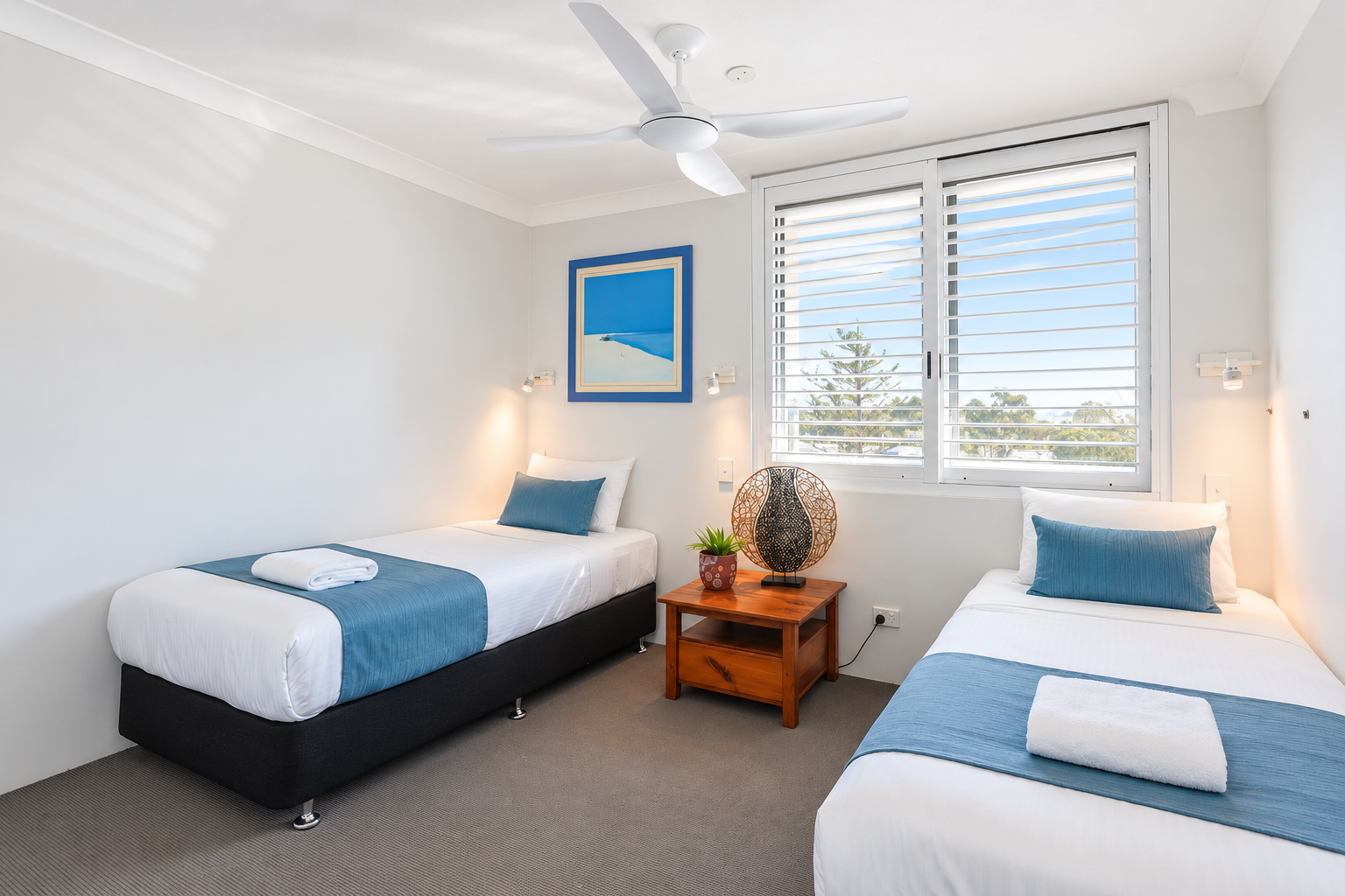Twin bedroom with two single beds featuring white linens and blue runners, a wooden nightstand in the center holding a decorative lamp and a small plant, white walls, a ceiling fan, and a window with white blinds showing trees outside.