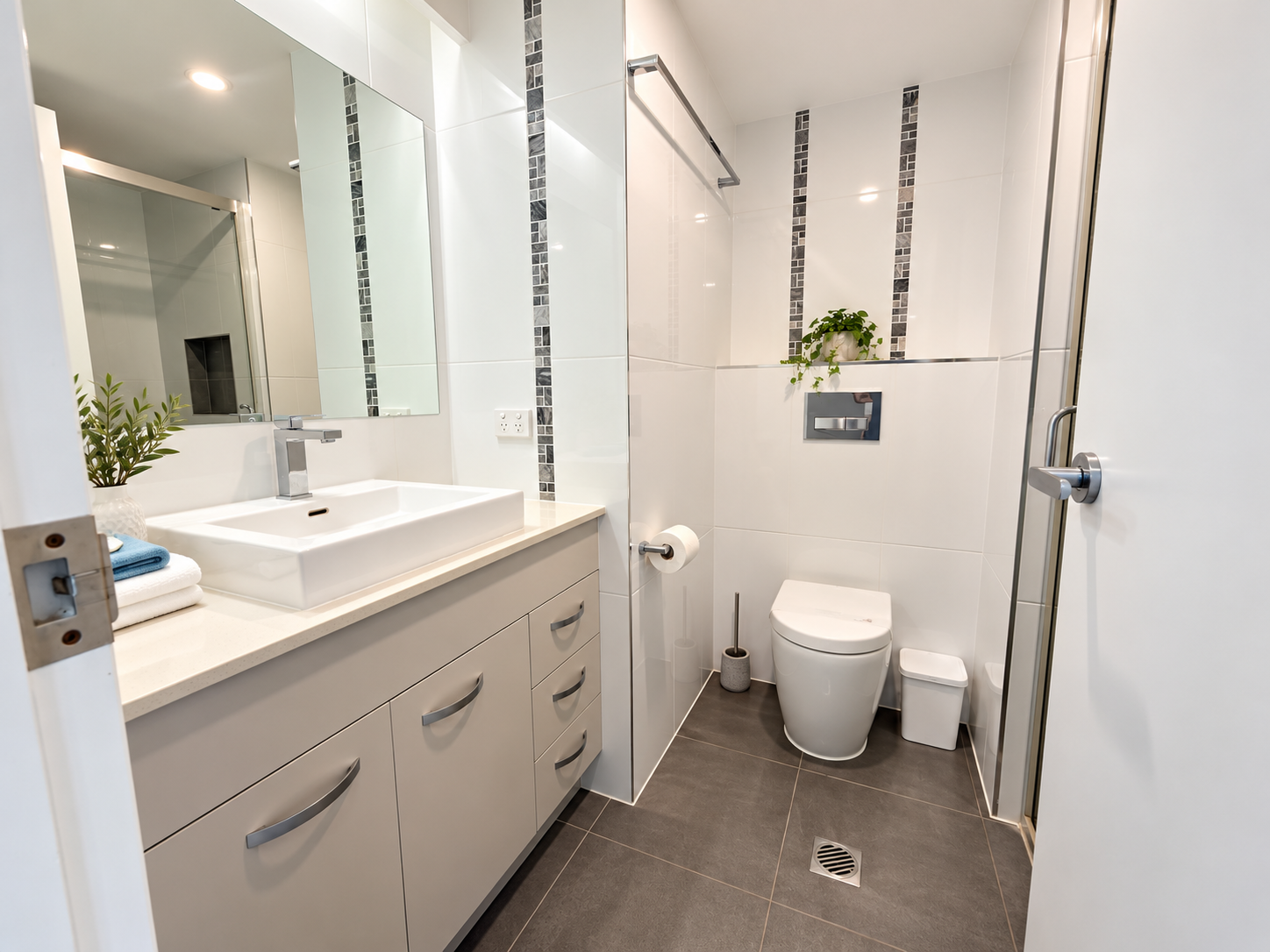 Modern bathroom with white vanity and rectangular sink on the left, large mirror above, and toilet with a plant on a tiled shelf on the right, white tiled walls with vertical dark mosaic tile accents, and dark gray floor tiles.
