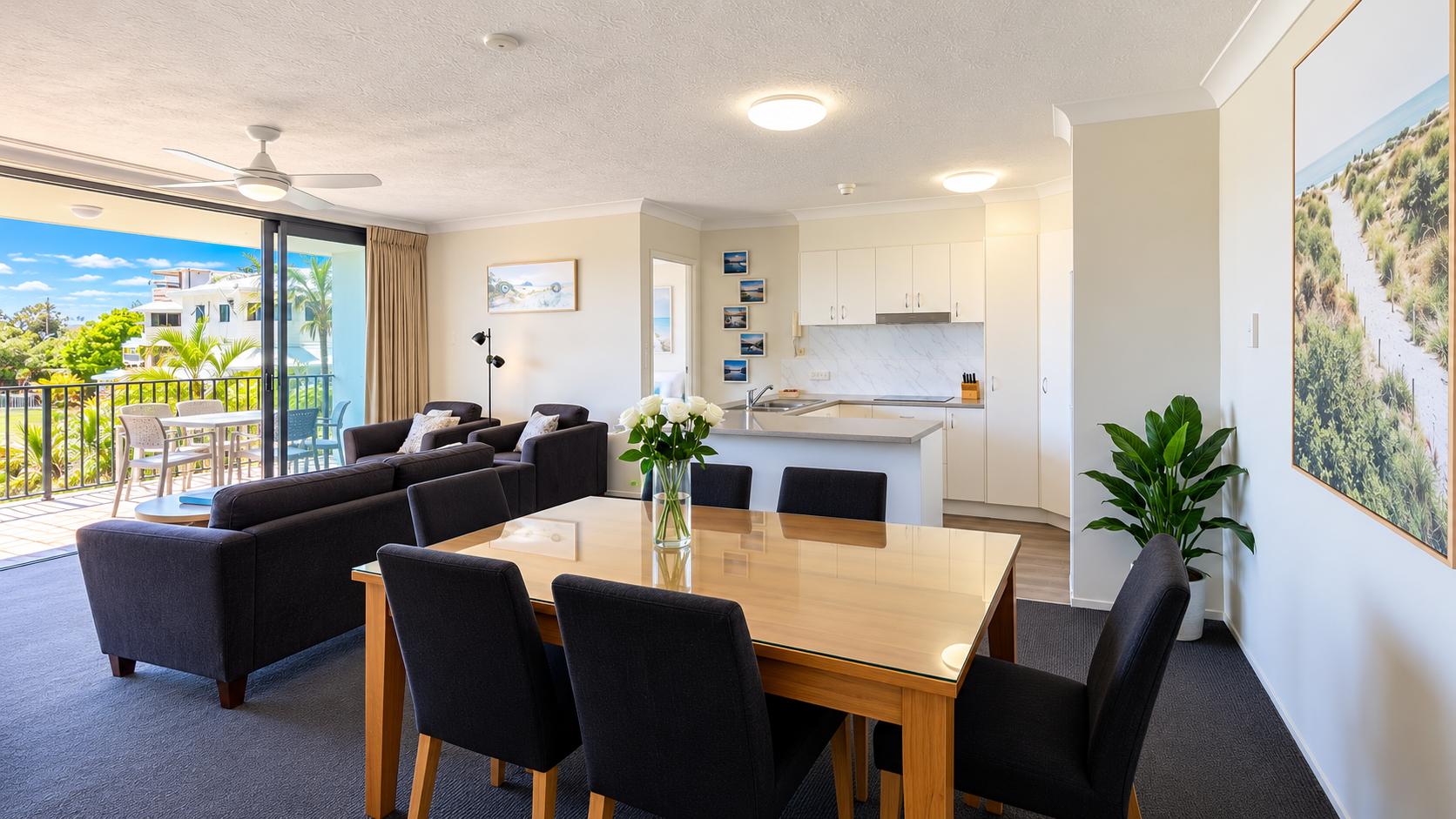 Modern living and dining area with a wooden table, six black chairs, and a vase of white roses, adjacent to a seating area with dark sofas and a glass coffee table, next to sliding glass doors opening to a balcony with outdoor furniture overlooking greenery and buildings, and an open kitchen with white cabinets and a marble backsplash.